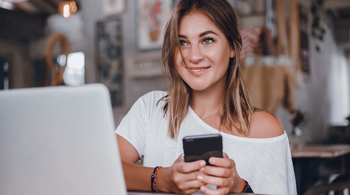 Young caucasian woman looks up from checking her mobile phone and browsing on a laptop