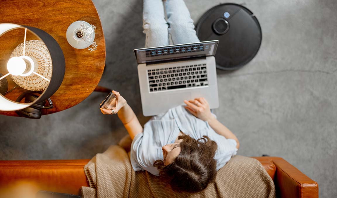 Aerial view of a caucasian woman sitting on the floor checking her smart phone while browsing on her laptop. Next to her, an automated robot vacuum cleans the floor