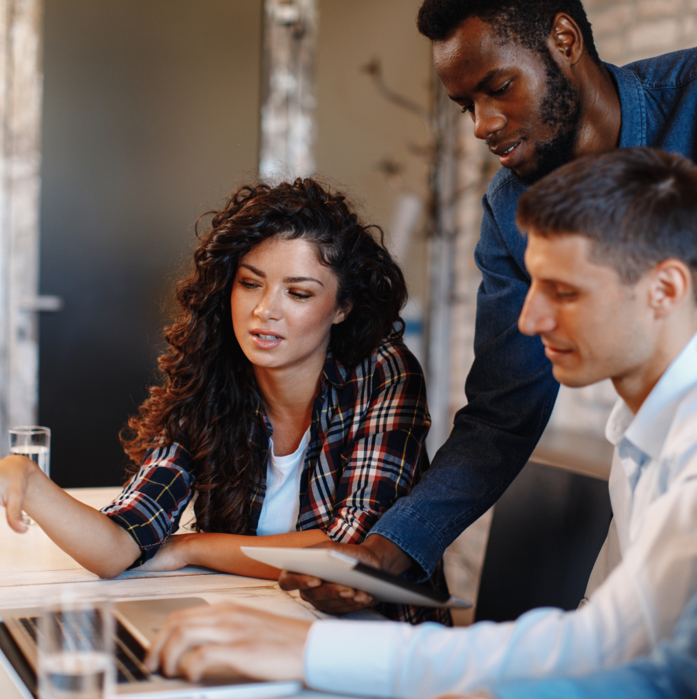 Three professionals collaborate at a wooden table, focused on a laptop. The woman on the left, with curly dark hair and a plaid shirt, gestures towards the screen, discussing a point. Next to her, a man with dark skin and wearing a dark blue shirt leans in to view the laptop, attentive to her explanation. On the right, another man in a light blue shirt also looks at the screen, involved in the conversation. The setting suggests a modern, informal office environment.