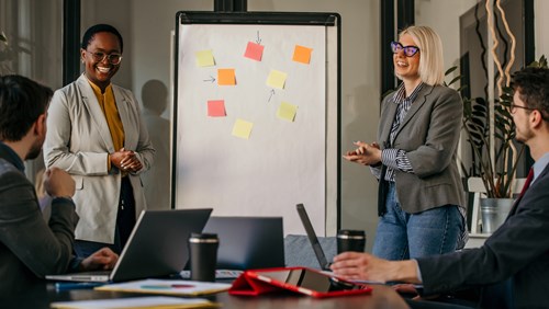 Two individuals in a professional setting, engaging in a discussion in front of a whiteboard covered with colorful sticky notes.
