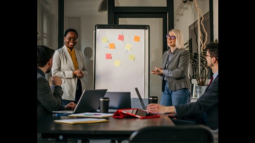 Two individuals in a professional setting, engaging in a discussion in front of a whiteboard covered with colorful sticky notes.