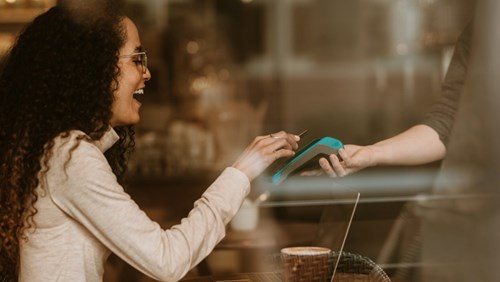 A smiling woman with curly hair holding her phone against a card reader to pay at a cafe, while holding a cup of coffee, with a laptop and notebook on the table