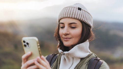 A woman in a knitted beanie and warm jacket, standing outdoors in a scenic area, smiles while looking at her smartphone
