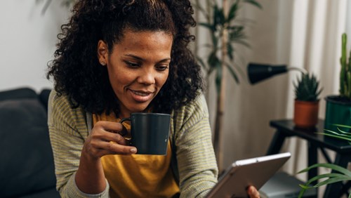 A woman with curly hair smiles while holding a black coffee mug and looking at a tablet screen in a cozy, plant-filled living room