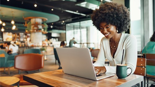 A woman with an afro hairstyle works on her laptop in a modern cafe, smiling with a cup of coffee on the table in front of her