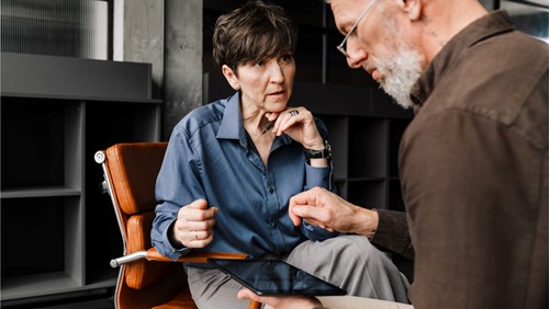 A man and a woman sit in an office, closely reviewing information on a tablet. The woman in a blue shirt listens intently as the man with gray hair explains something