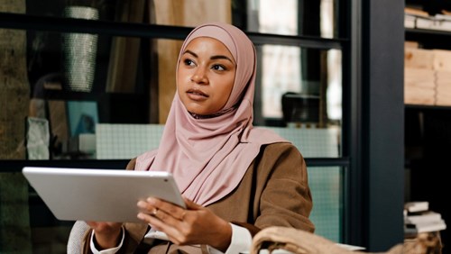 A woman wearing a light pink hijab and a brown jacket is holding a tablet while sitting in a modern indoor setting. She appears thoughtful, gazing into the distance, with shelves and a large window visible in the background. The scene conveys a professional and focused atmosphere.