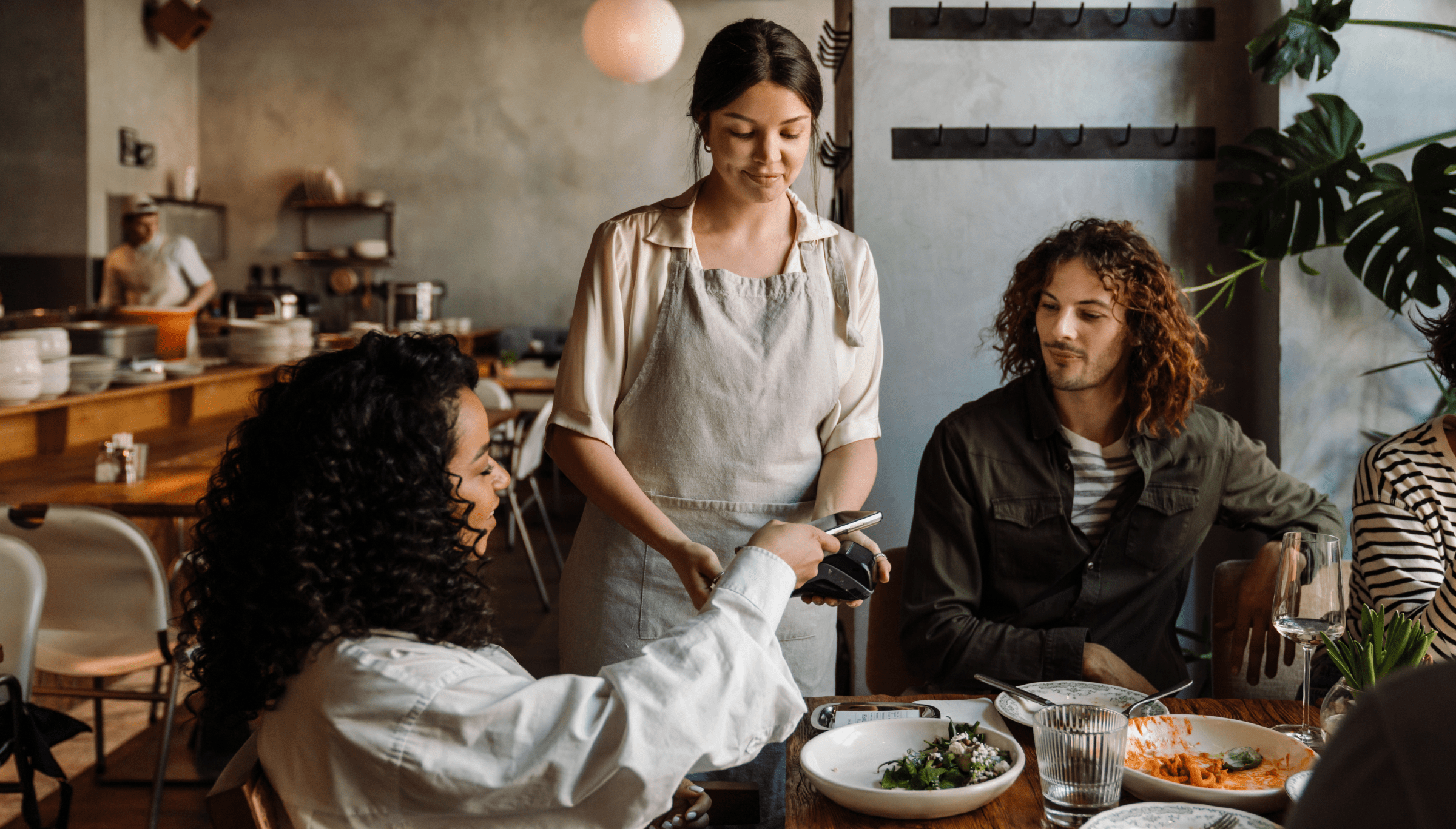 A group of diners at a restaurant are paying for their meal. One woman, seated at the table, is holding a smartphone and using a contactless payment system held by the server, who is wearing an apron. Another person at the table looks on, smiling. The setting is a warm, cozy restaurant with wooden tables and a relaxed atmosphere, where people are enjoying their meal. The background shows additional diners and kitchen staff, contributing to the bustling yet comfortable ambiance.