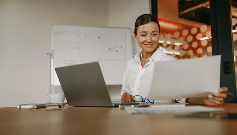 A woman is seated at a desk, smiling as she reviews a document in her hand. She has a laptop open in front of her, along with a cup of coffee, a notebook, and a pair of glasses resting on the table. Behind her, a whiteboard with diagrams is visible, and the background features a modern office setting with red and white polka-dot accents. The scene conveys a professional and positive work environment, with the woman appearing engaged and content with her work.