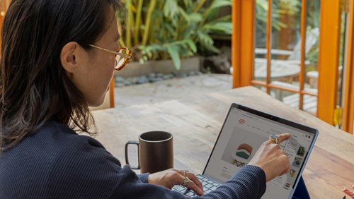 A person wearing glasses is sitting at a wooden table, using a tablet with a keyboard attachment. They are pointing at something on the screen, which displays a webpage with images of products, possibly on an online shopping site. A black coffee mug is placed next to them on the table. The setting is bright and airy, with large glass doors opening to a garden or outdoor area with lush greenery, creating a serene and focused work environment.