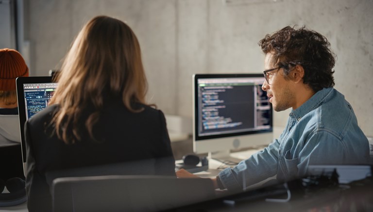 Two people are working on coding projects in a modern office environment. They are seated side by side, each with a computer screen displaying lines of code. The person on the right, wearing glasses and a denim shirt, is engaged in a discussion or collaboration with the person next to them, who is partially turned away from the camera. The setting suggests a focused and collaborative work atmosphere, with both individuals deeply involved in software development or programming tasks.