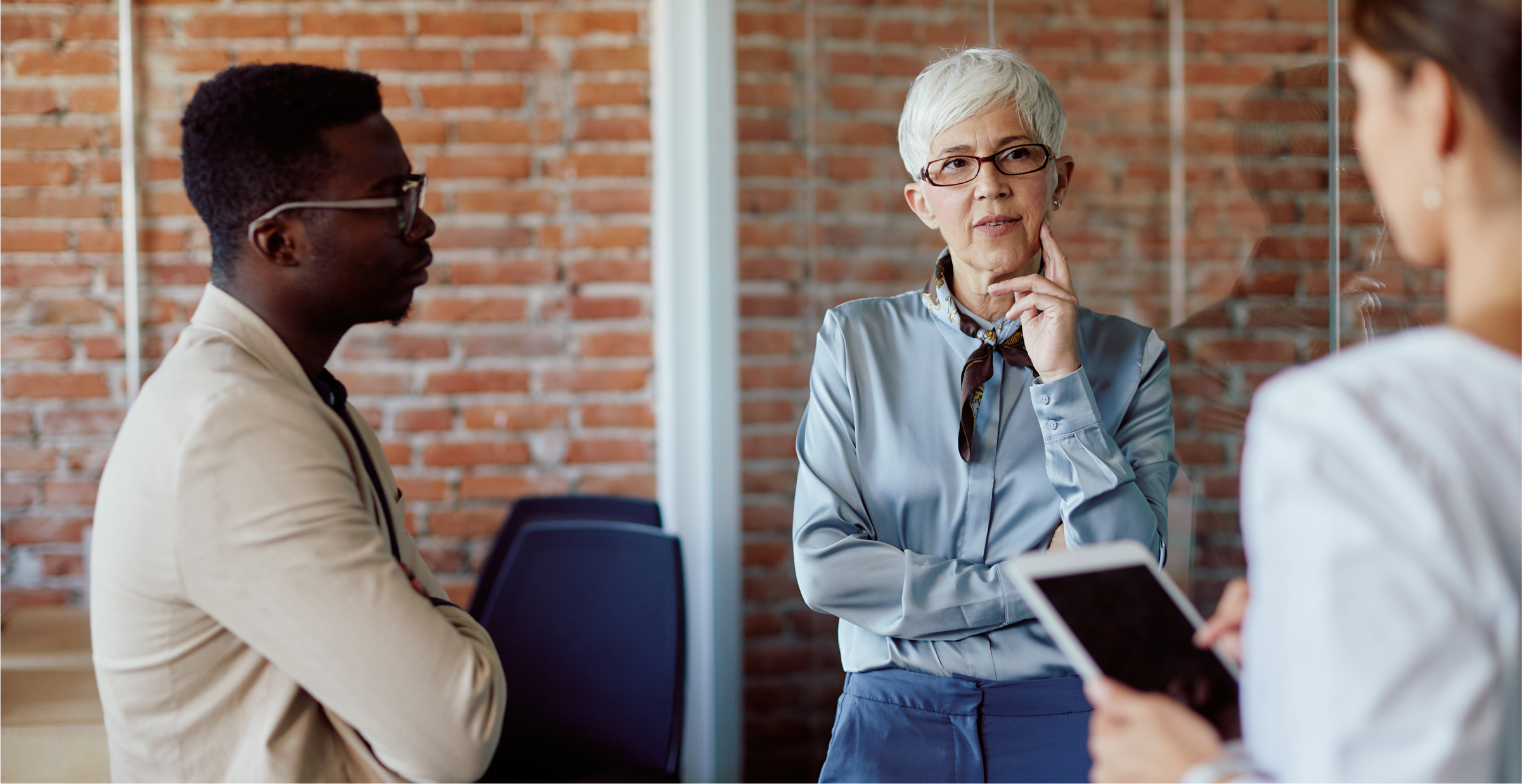 A diverse group of professionals engaged in a thoughtful discussion in a modern office setting. The group includes a senior woman with short white hair and glasses, who is speaking while holding her chin, and a younger man listening intently with his arms crossed. Another person, partially visible and holding a tablet, is involved in the conversation. The background features an exposed brick wall and glass partitions, giving the office a contemporary, open feel. The atmosphere suggests a strategic meeting or brainstorming session.