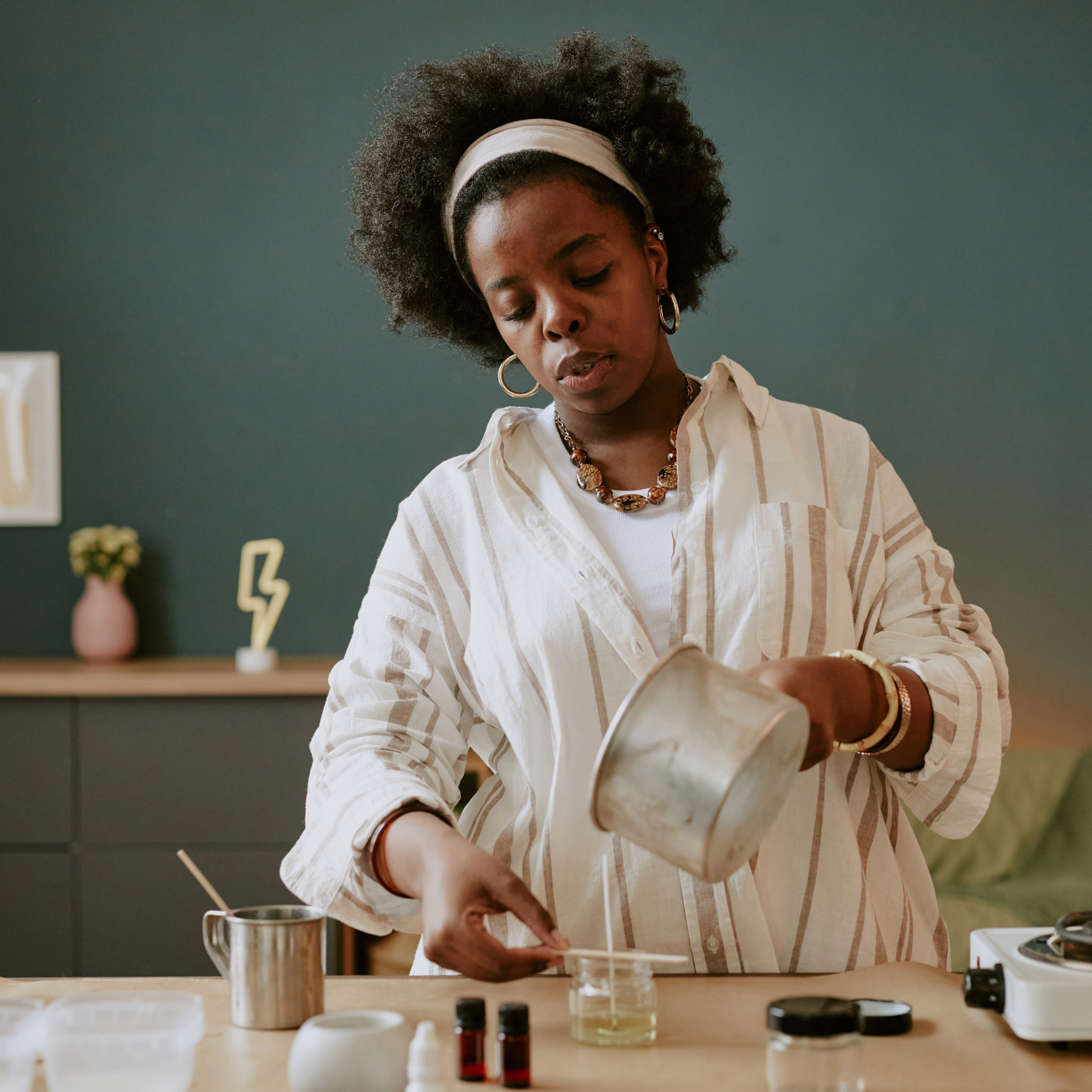 A woman with a headband and natural hair is carefully pouring liquid wax into a container, likely making a candle