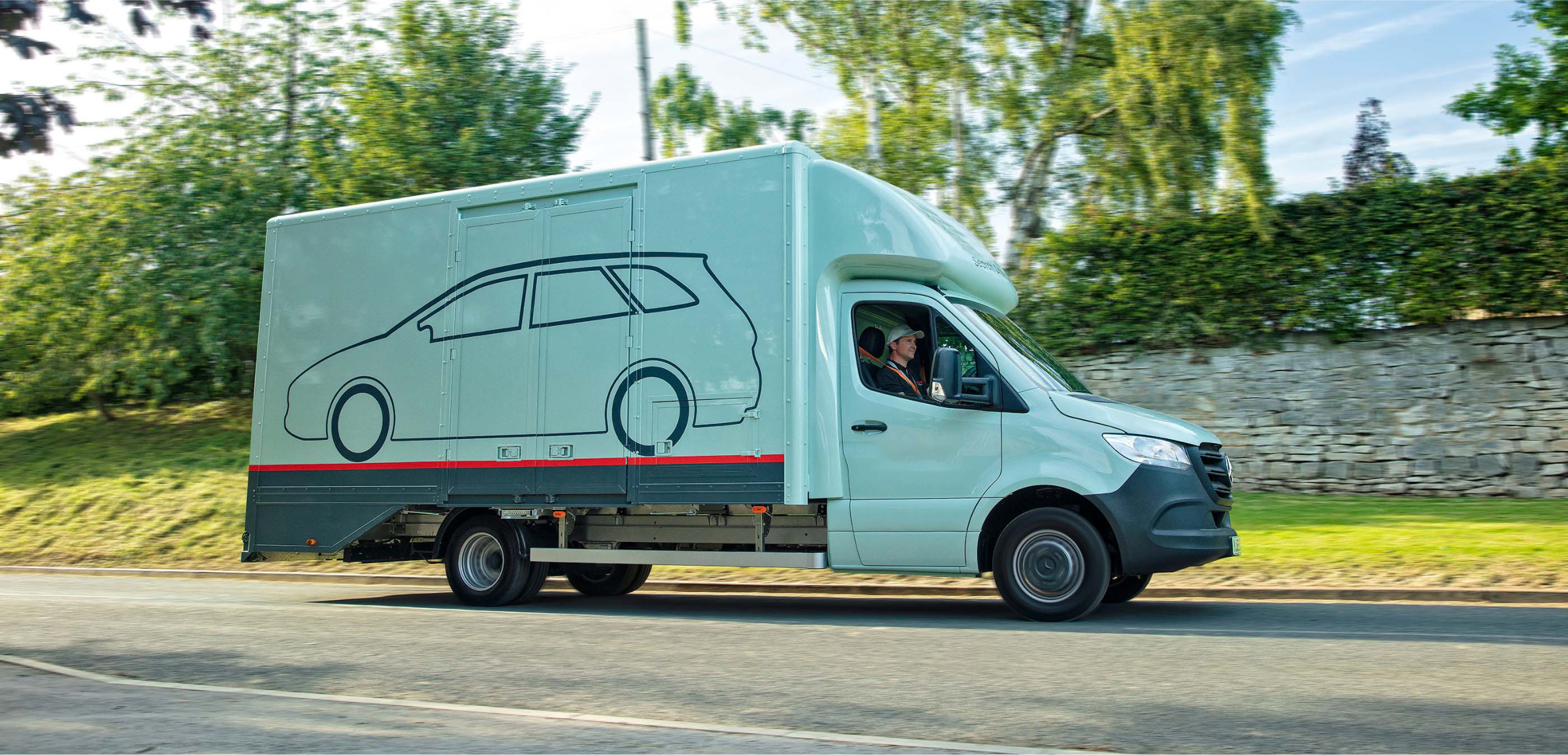 A large delivery van is driving down a suburban street, with the driver visible and focused on the road.