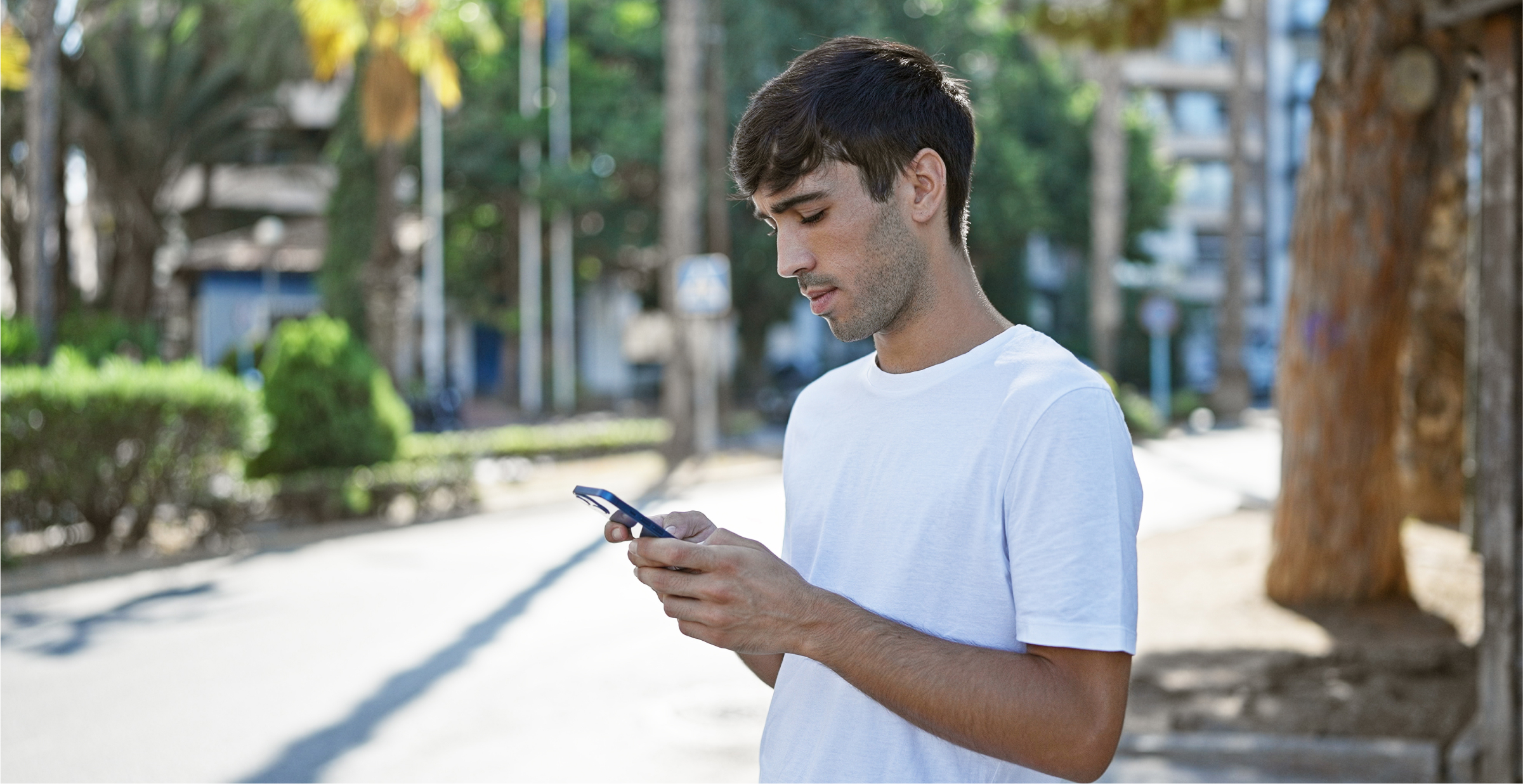 A young man wearing a white t-shirt is standing outdoors on a sunny day, looking down at his smartphone. He appears focused on the screen, possibly reading a message or navigating. The background shows a tree-lined street with some buildings and greenery, suggesting an urban setting. The lighting is bright, indicating that the photo was taken during daylight hours.