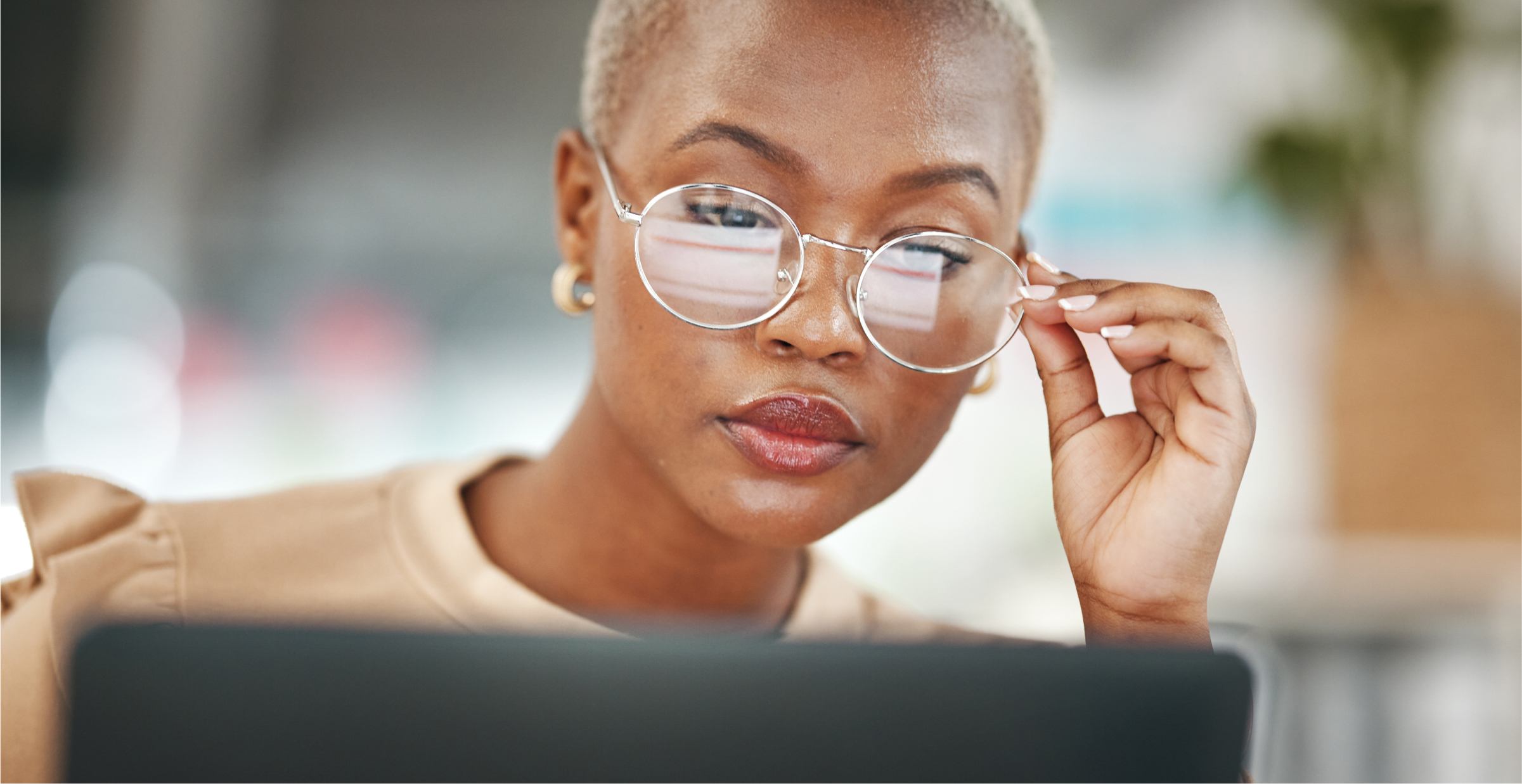 A close-up of a woman with short hair and large, round glasses, intently focused on her work. She is adjusting her glasses while looking at a computer screen, reflecting concentration and determination. The background is softly blurred, keeping the attention on her thoughtful expression and professional demeanor. The scene conveys a sense of focus and dedication in a modern, possibly office or remote work environment.