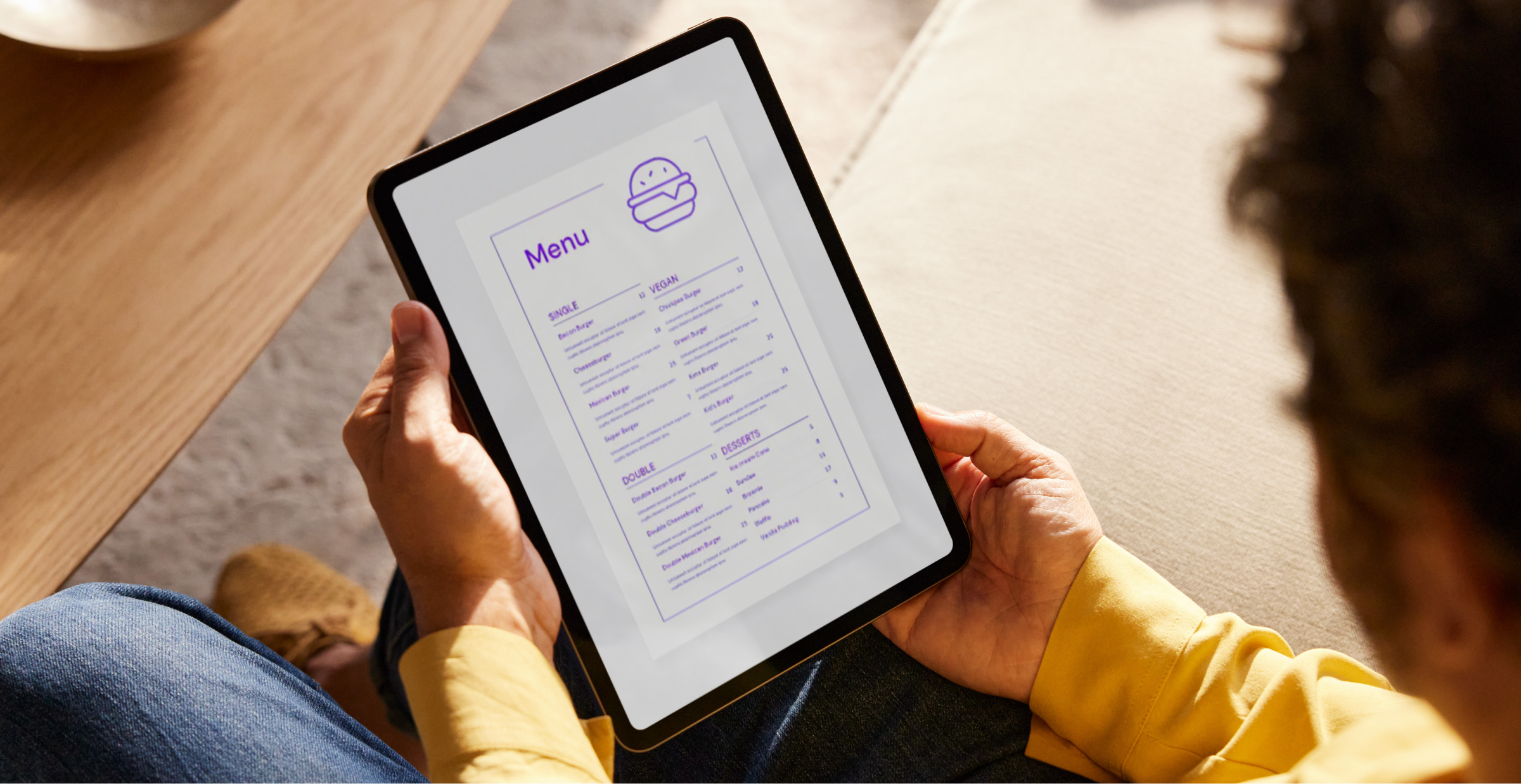 A person is holding a tablet, looking at a digital menu displayed on the screen. The menu features various food items, including vegan and dessert options, with a simple, clean design in purple text. The person is seated in a comfortable indoor setting, with a wooden table and a cozy environment visible in the background. The scene suggests the use of technology for ordering food or browsing restaurant options, highlighting the convenience and modern approach to dining.
