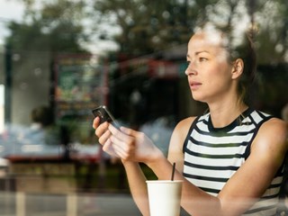 Woman using a smartphone in a cafe, seen through a glass window, with a contemplative expression.