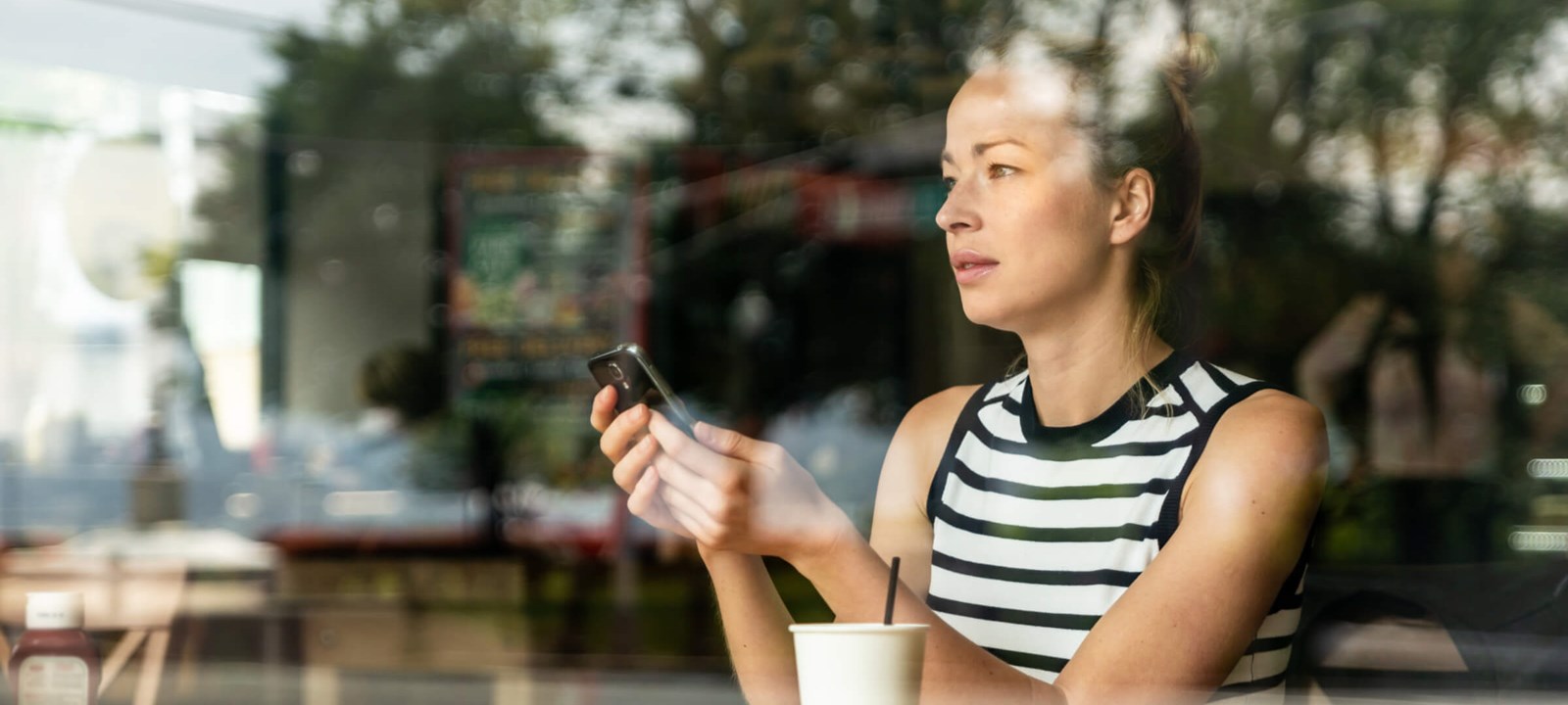 Woman using a smartphone in a cafe, seen through a glass window, with a contemplative expression.