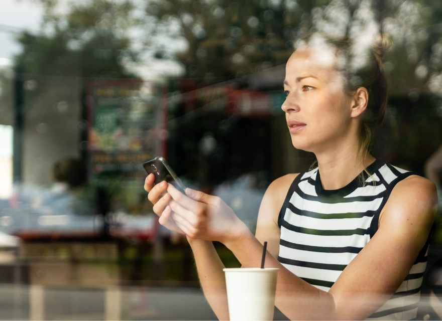 Woman using a smartphone in a cafe, seen through a glass window, with a contemplative expression.