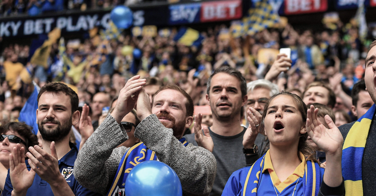 Football fans enthusiastically cheering in the stands, some clapping and others shouting in support, with a sea of yellow and blue colors around them, indicating strong team spirit and unity.