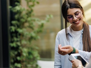 A smiling woman wearing glasses and a light blue shirt is using her smartwatch to make a contactless payment. She holds her wrist near a payment terminal, and the scene takes place in a bright, modern outdoor setting with greenery in the background. The image conveys a sense of convenience and technology in everyday life, highlighting the ease of digital payments through wearable devices.