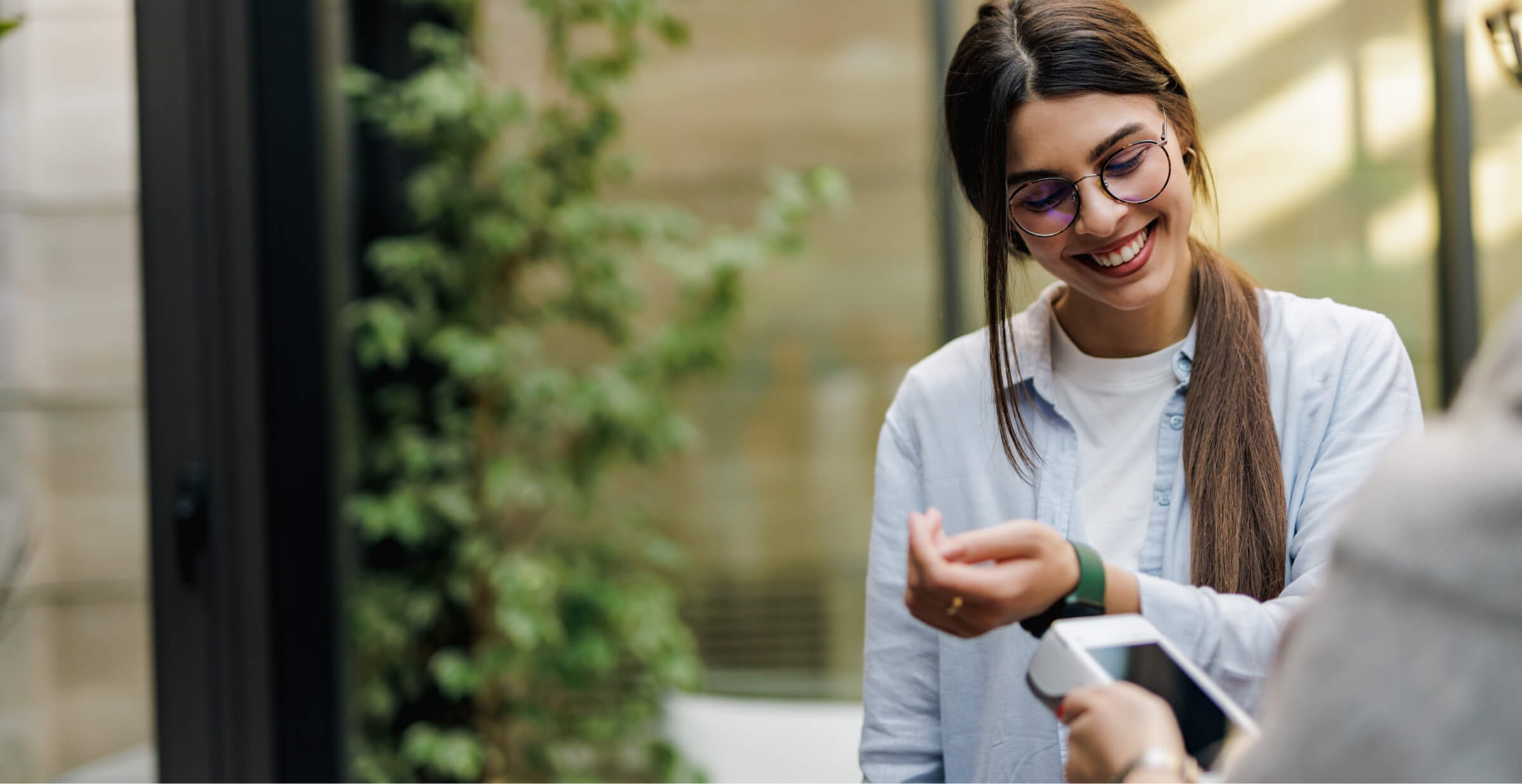 A smiling woman wearing glasses and a light blue shirt is using her smartwatch to make a contactless payment. She holds her wrist near a payment terminal, and the scene takes place in a bright, modern outdoor setting with greenery in the background. The image conveys a sense of convenience and technology in everyday life, highlighting the ease of digital payments through wearable devices.