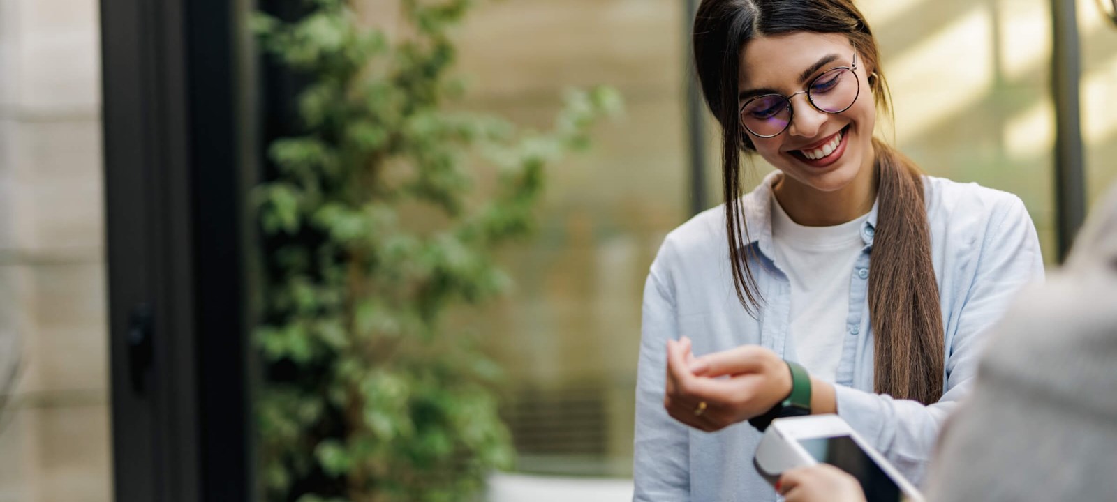 A smiling woman wearing glasses and a light blue shirt is using her smartwatch to make a contactless payment. She holds her wrist near a payment terminal, and the scene takes place in a bright, modern outdoor setting with greenery in the background. The image conveys a sense of convenience and technology in everyday life, highlighting the ease of digital payments through wearable devices.
