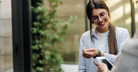 A smiling woman wearing glasses and a light blue shirt is using her smartwatch to make a contactless payment. She holds her wrist near a payment terminal, and the scene takes place in a bright, modern outdoor setting with greenery in the background. The image conveys a sense of convenience and technology in everyday life, highlighting the ease of digital payments through wearable devices.