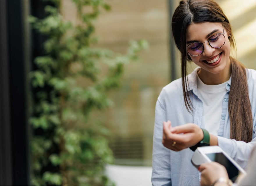 A smiling woman wearing glasses and a light blue shirt is using her smartwatch to make a contactless payment. She holds her wrist near a payment terminal, and the scene takes place in a bright, modern outdoor setting with greenery in the background. The image conveys a sense of convenience and technology in everyday life, highlighting the ease of digital payments through wearable devices.