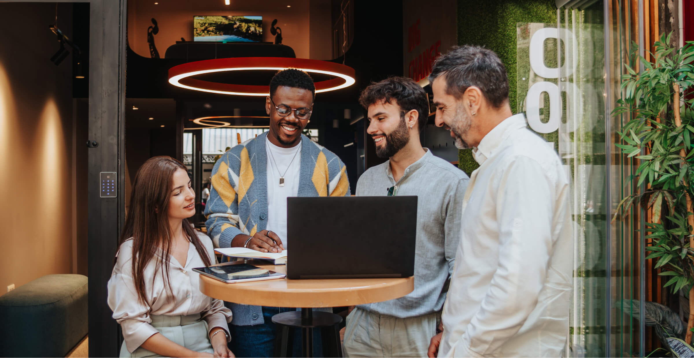 A diverse group of colleagues is gathered around a laptop during an outdoor meeting. The group consists of four men and one woman, all smiling and engaged in a collaborative discussion. They are standing and sitting around a high table in a modern outdoor space, with greenery and contemporary architecture in the background. The setting is casual and vibrant, reflecting a positive and creative work environment.