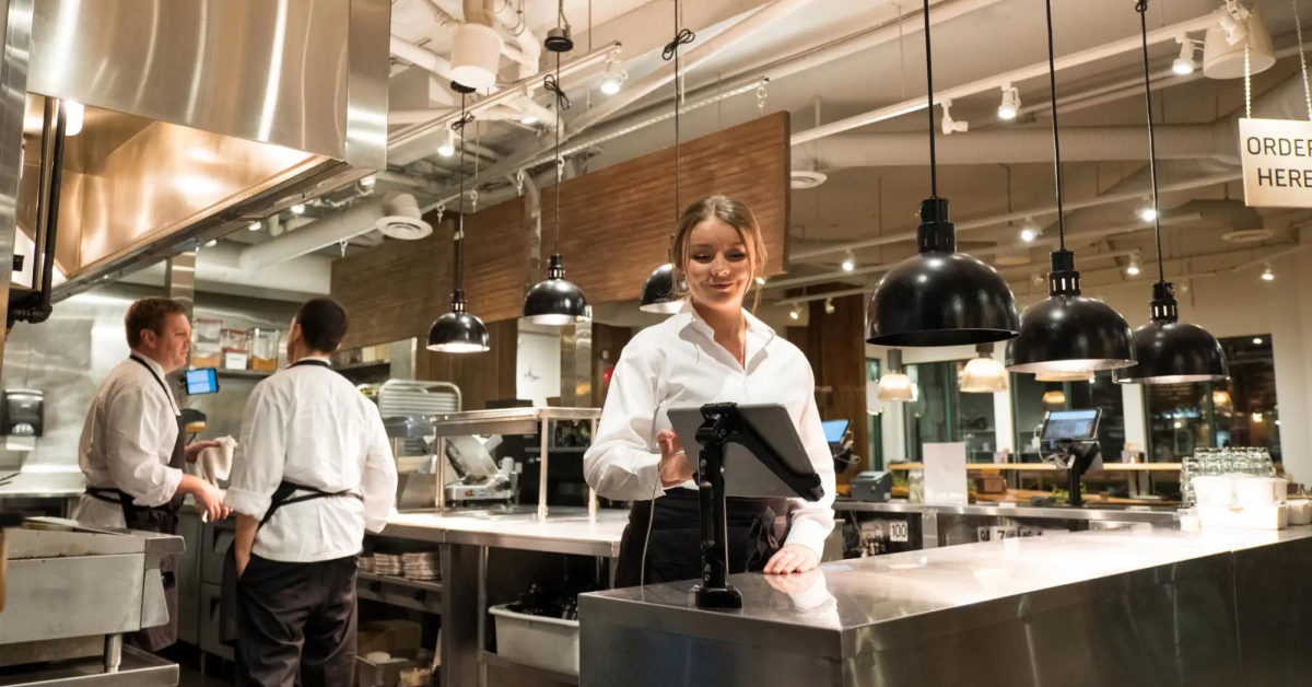 A server in a white shirt standing in a modern, well-lit kitchen, smiling as she interacts with a mounted tablet. The kitchen features industrial-style lighting and stainless steel surfaces, with another staff member working in the background.