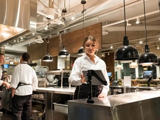 A server in a white shirt standing in a modern, well-lit kitchen, smiling as she interacts with a mounted tablet. The kitchen features industrial-style lighting and stainless steel surfaces, with another staff member working in the background.