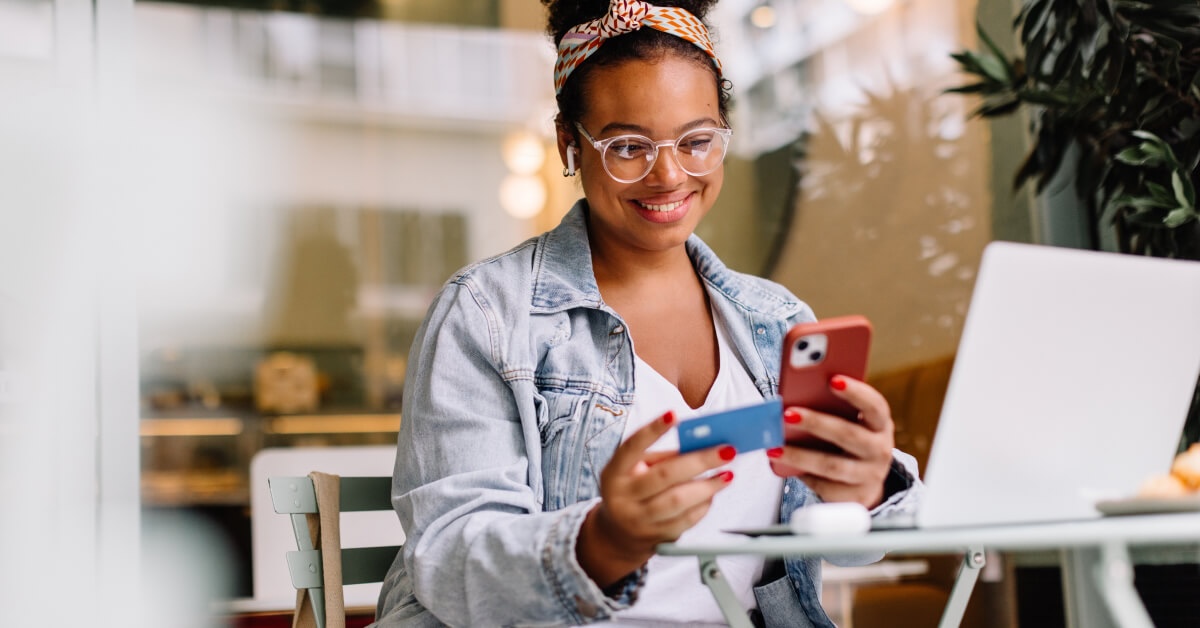 A young woman sitting at a cafe table, smiling as she uses her smartphone and holds a credit card, with a laptop open in front of her. She is wearing a denim jacket, glasses, and a colorful headband, suggesting she is making an online purchase or transaction in a relaxed setting.