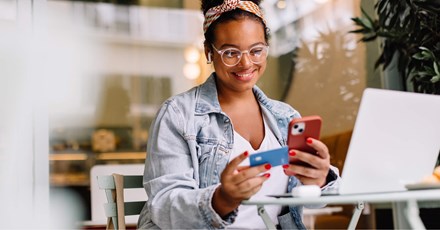 A young woman sitting at a cafe table, smiling as she uses her smartphone and holds a credit card, with a laptop open in front of her. She is wearing a denim jacket, glasses, and a colorful headband, suggesting she is making an online purchase or transaction in a relaxed setting.