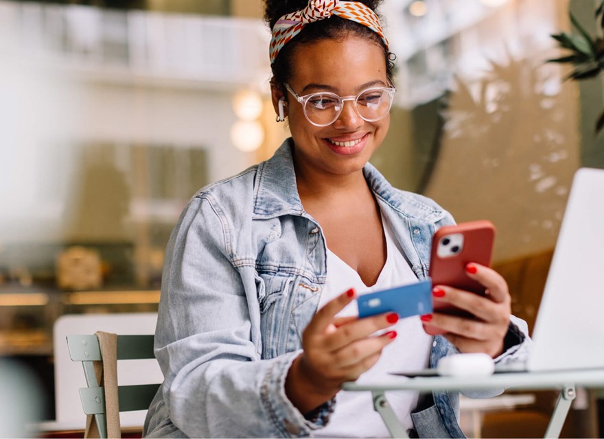 A young woman sitting at a cafe table, smiling as she uses her smartphone and holds a credit card, with a laptop open in front of her. She is wearing a denim jacket, glasses, and a colorful headband, suggesting she is making an online purchase or transaction in a relaxed setting.