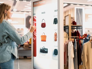 Woman using an interactive digital kiosk to browse handbags in a modern retail store.