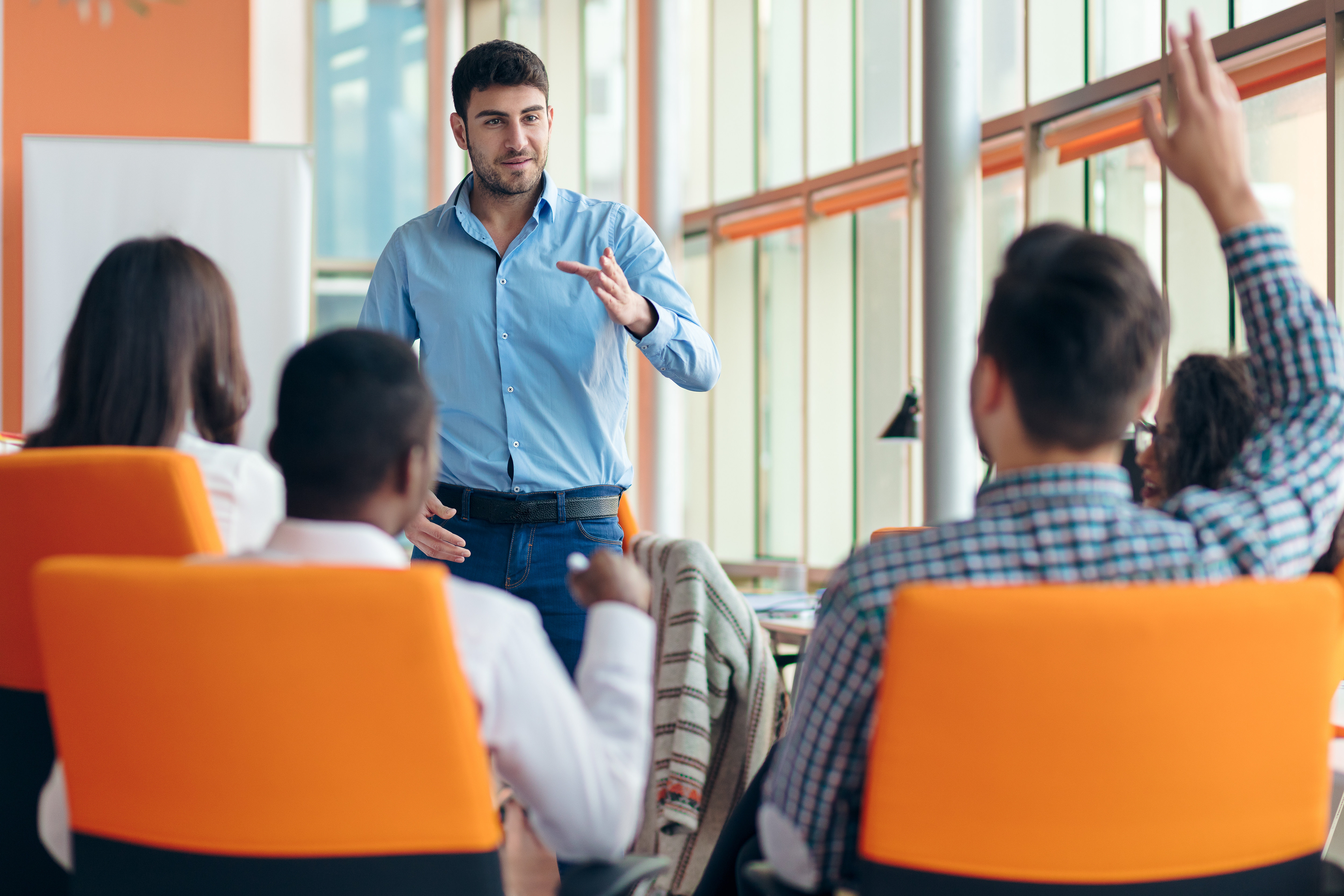 A group of people on orange chairs. A man has his hand raised.