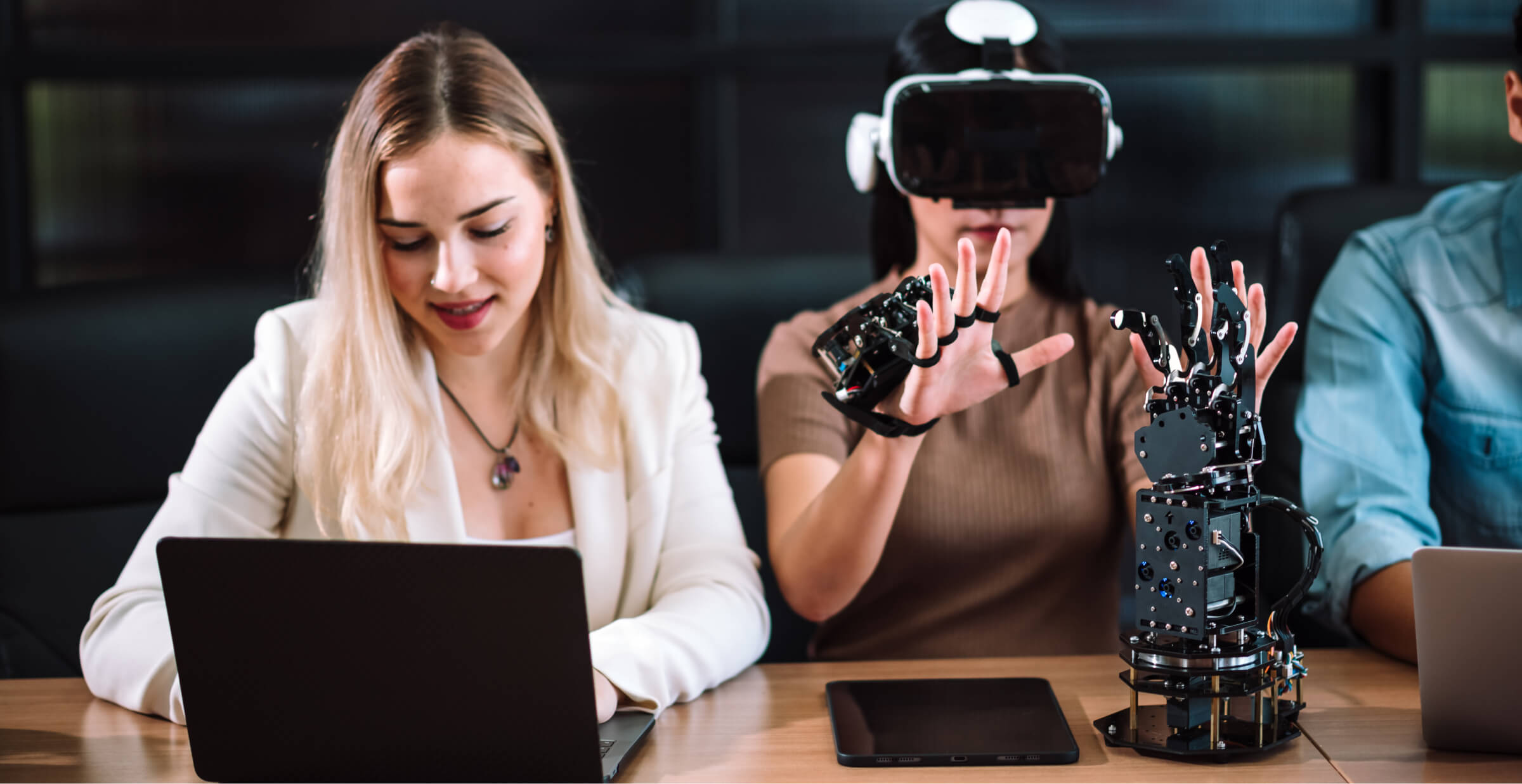 Woman using VR gloves and headset for robotics testing, while colleague works on laptop.