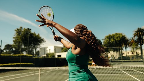 A woman with braided hair is playing tennis on an outdoor court. She is in mid-motion, preparing to serve the ball with her racket. The sun is shining brightly, and the sky is clear, creating a vibrant and energetic atmosphere. The background shows lush greenery and buildings, adding to the picturesque setting. The image captures the athleticism and focus of the tennis player.