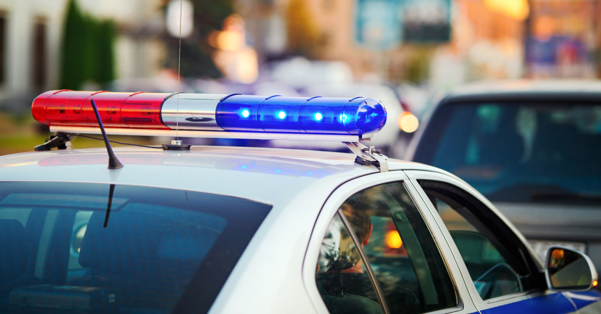 A close-up of a police car’s roof with its red and blue lights flashing. The background shows a blurred city street with other vehicles, suggesting the police car is responding to an incident or patrolling in an urban area. The image captures the sense of urgency and authority associated with law enforcement in action.