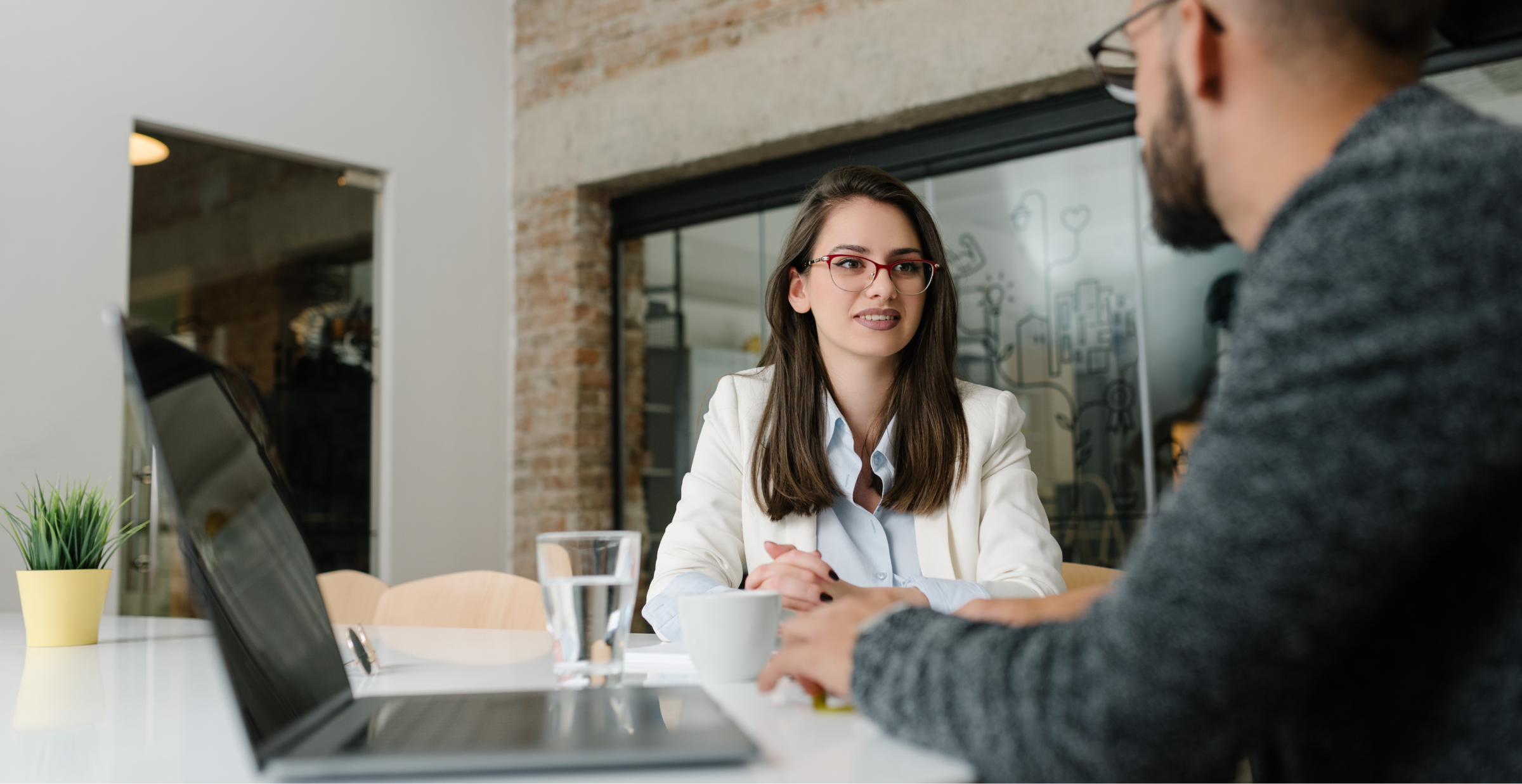 Two people are sitting at a table in an office setting, engaged in conversation. One person is using a laptop and facing the other, who is wearing glasses and a white blazer. A glass of water and a potted plant are on the table.