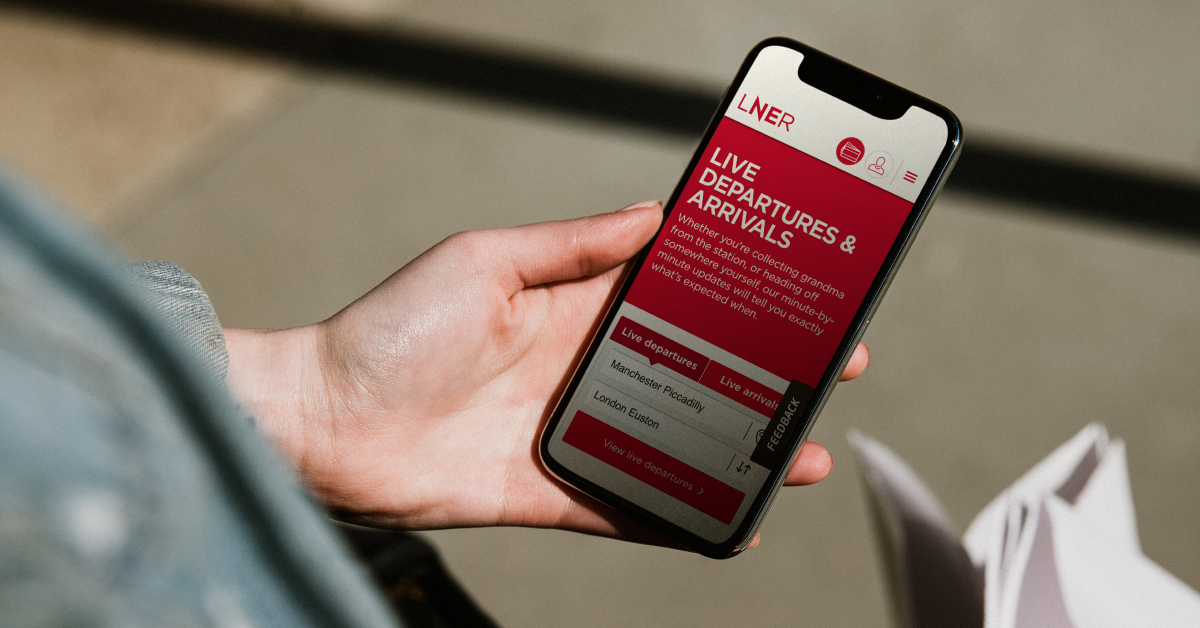 A person holding a smartphone displaying the LNER app, which shows live departures and arrivals information for train stations, with a focus on Manchester Piccadilly and London Euston.