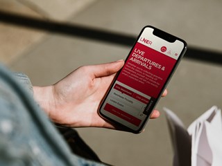 A person holding a smartphone displaying the LNER app, which shows live departures and arrivals information for train stations, with a focus on Manchester Piccadilly and London Euston.