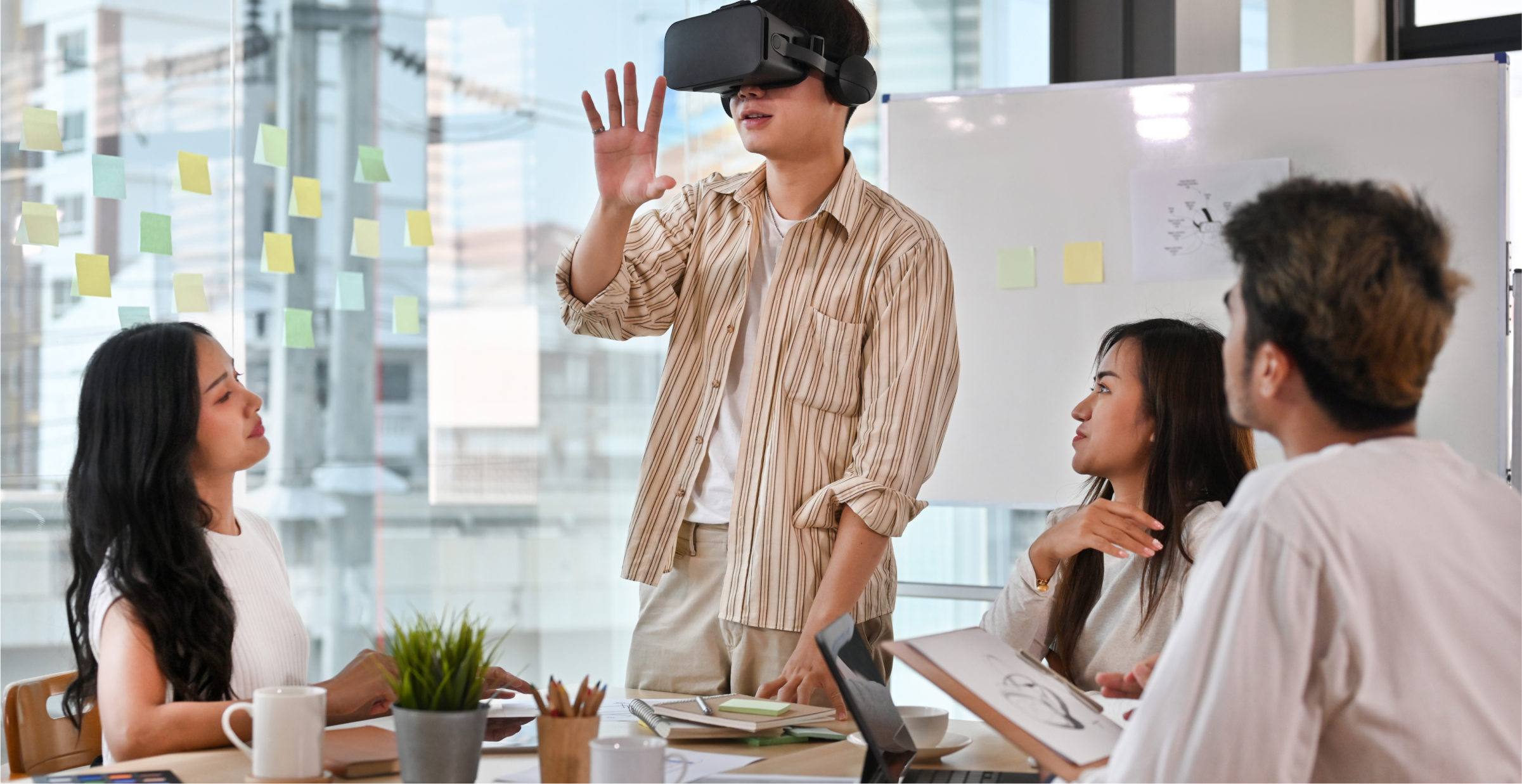 A group of four colleagues in a modern office participate in a team meeting. One person, standing, is wearing a virtual reality (VR) headset and gesturing with their hand as they demonstrate or interact with the VR environment. The other three team members are seated around a table, observing and discussing the presentation. The room features large windows with sticky notes on the glass, a whiteboard in the background, and a mix of notebooks, coffee cups, and a small potted plant on the table, creating a collaborative and innovative atmosphere.