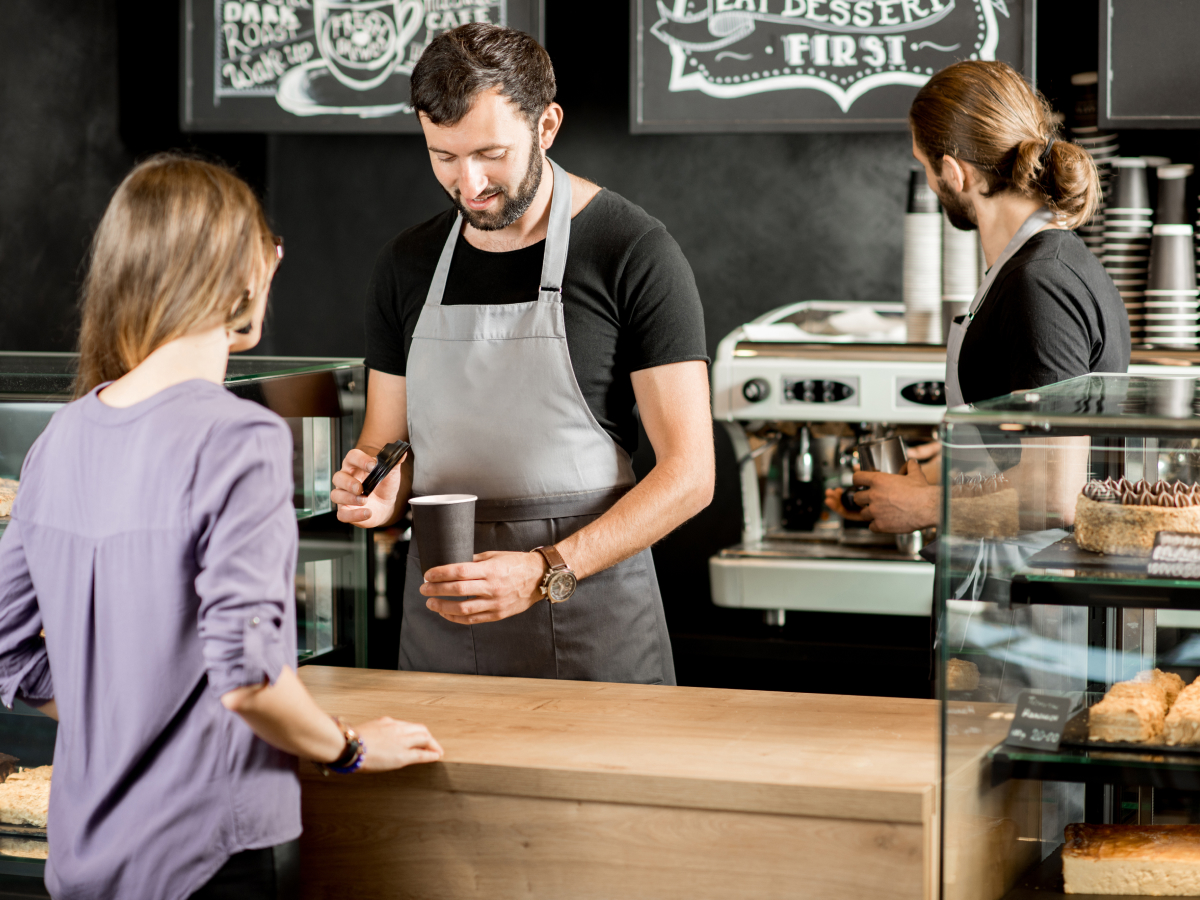 A barista in a gray apron preparing a coffee order for a customer at a modern coffee shop. The customer stands at the counter, while another barista works in the background near an espresso machine. The shop has a cozy, artisanal atmosphere with chalkboard menus in the background.
