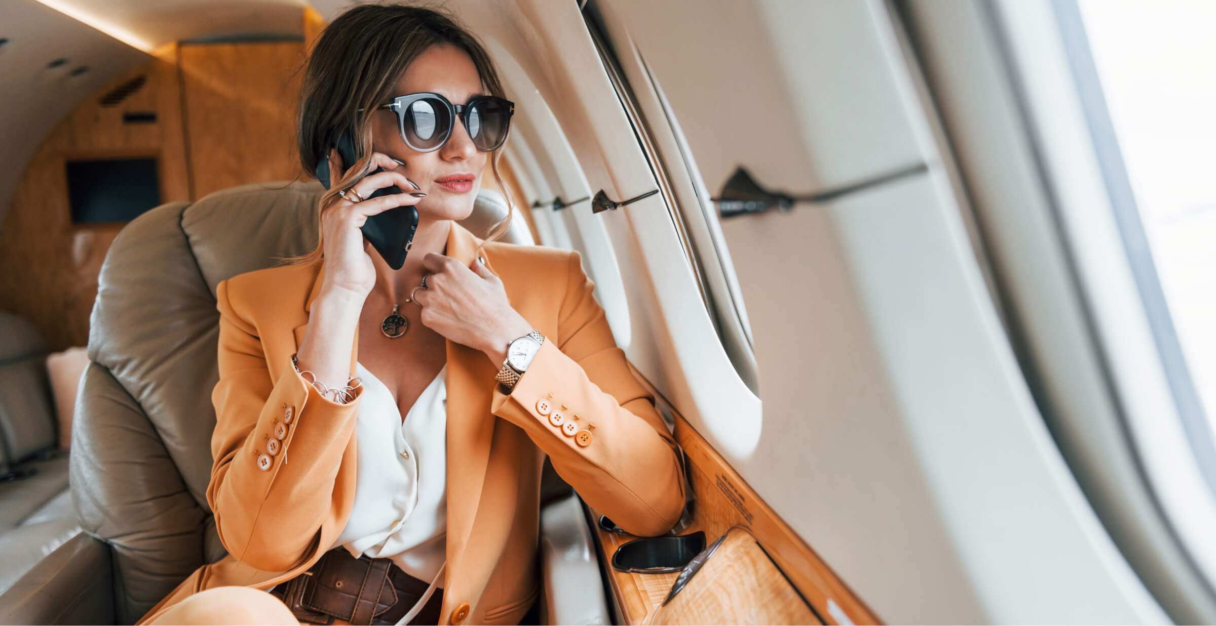 A stylish businesswoman wearing a light orange suit and large sunglasses is sitting in a private jet, talking on her phone. She looks composed and confident, with the luxurious interior of the jet visible around her. The image captures a moment of professional engagement during travel, highlighting themes of success, luxury, and mobility in a business context.
