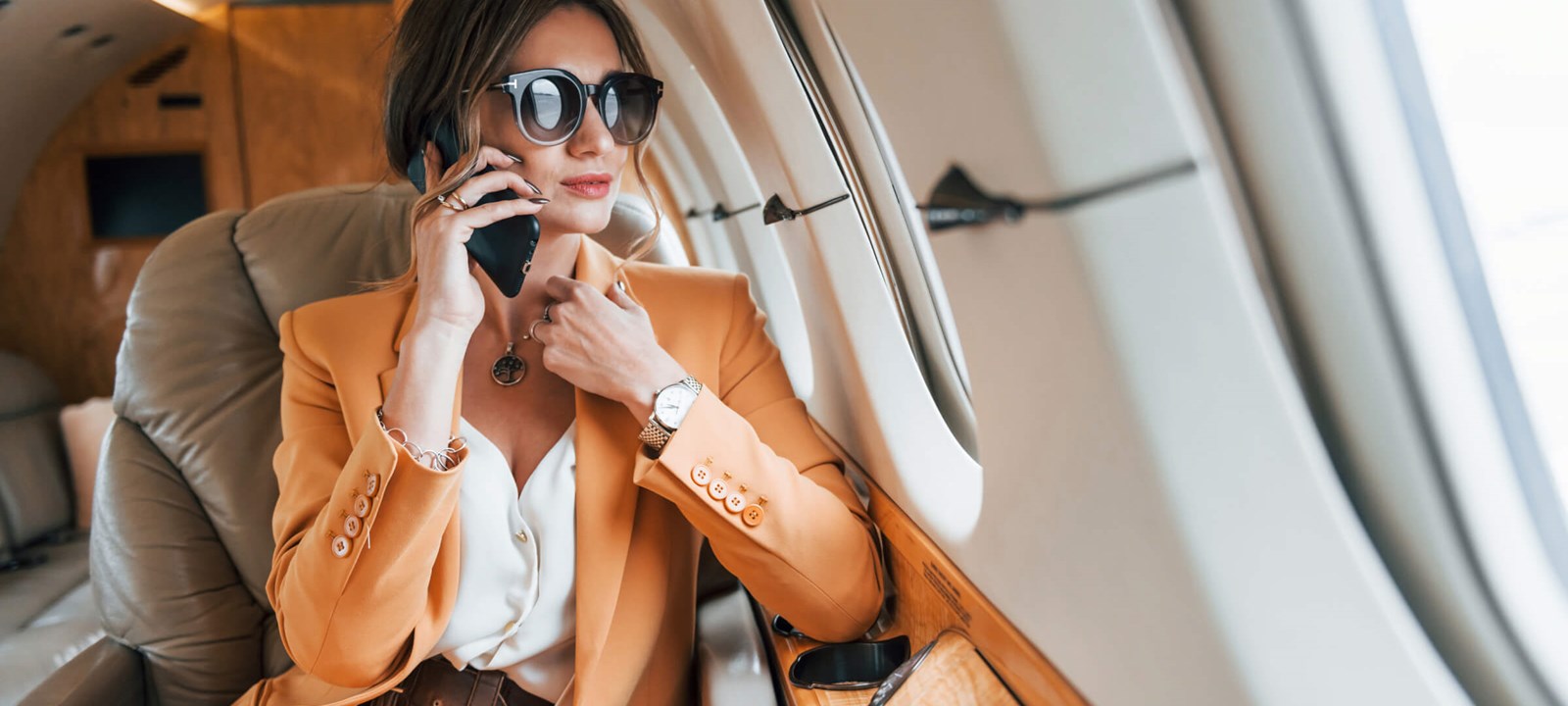 A stylish businesswoman wearing a light orange suit and large sunglasses is sitting in a private jet, talking on her phone. She looks composed and confident, with the luxurious interior of the jet visible around her. The image captures a moment of professional engagement during travel, highlighting themes of success, luxury, and mobility in a business context.