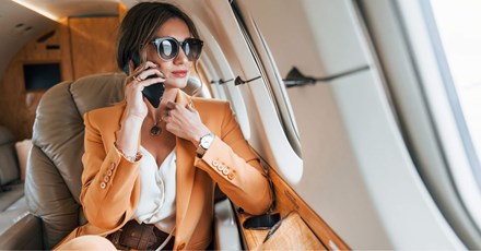 A stylish businesswoman wearing a light orange suit and large sunglasses is sitting in a private jet, talking on her phone. She looks composed and confident, with the luxurious interior of the jet visible around her. The image captures a moment of professional engagement during travel, highlighting themes of success, luxury, and mobility in a business context.