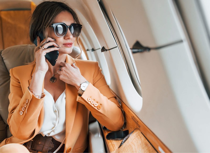 A stylish businesswoman wearing a light orange suit and large sunglasses is sitting in a private jet, talking on her phone. She looks composed and confident, with the luxurious interior of the jet visible around her. The image captures a moment of professional engagement during travel, highlighting themes of success, luxury, and mobility in a business context.